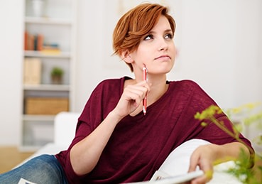 Woman sitting with a pen on her couch pondering a question. 